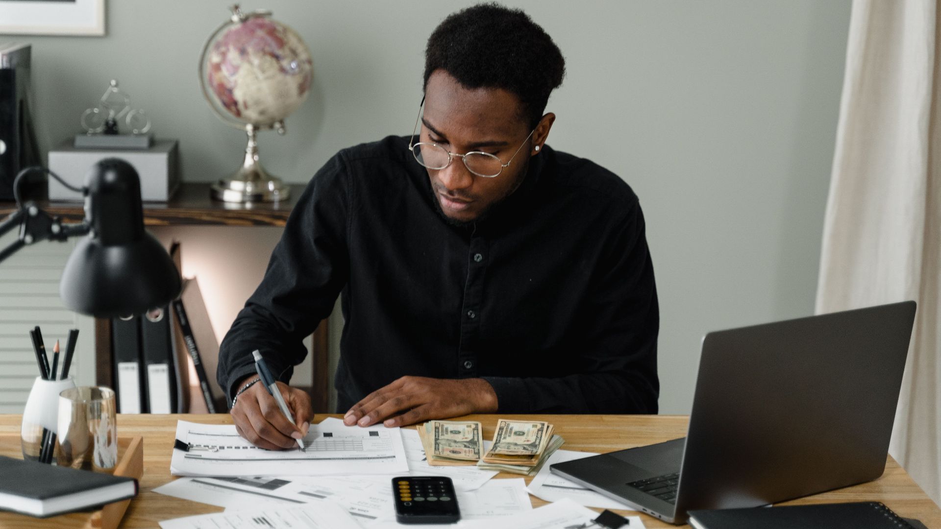 An adult man calculates expenses, using a laptop and documents at a desk in a home office setting.