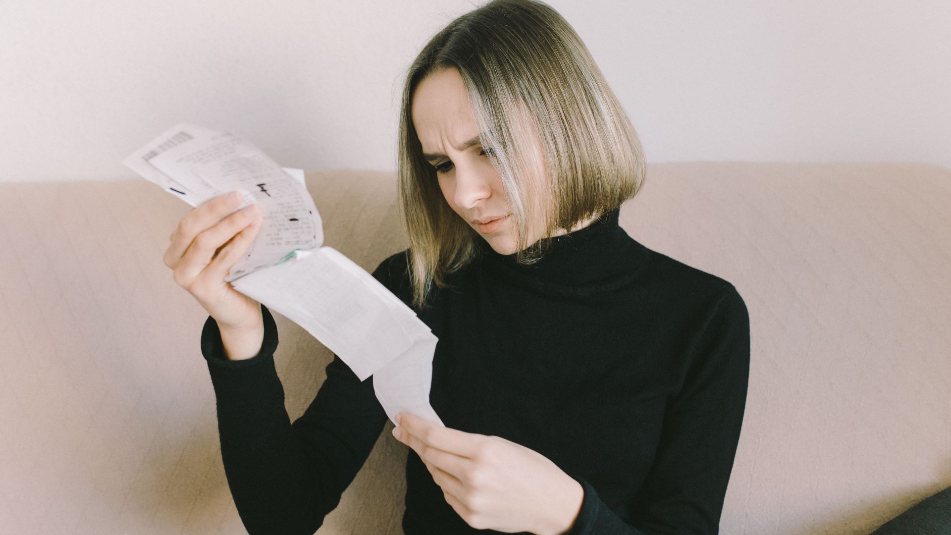 Concerned woman in black sweater examining bills on beige sofa indoors.