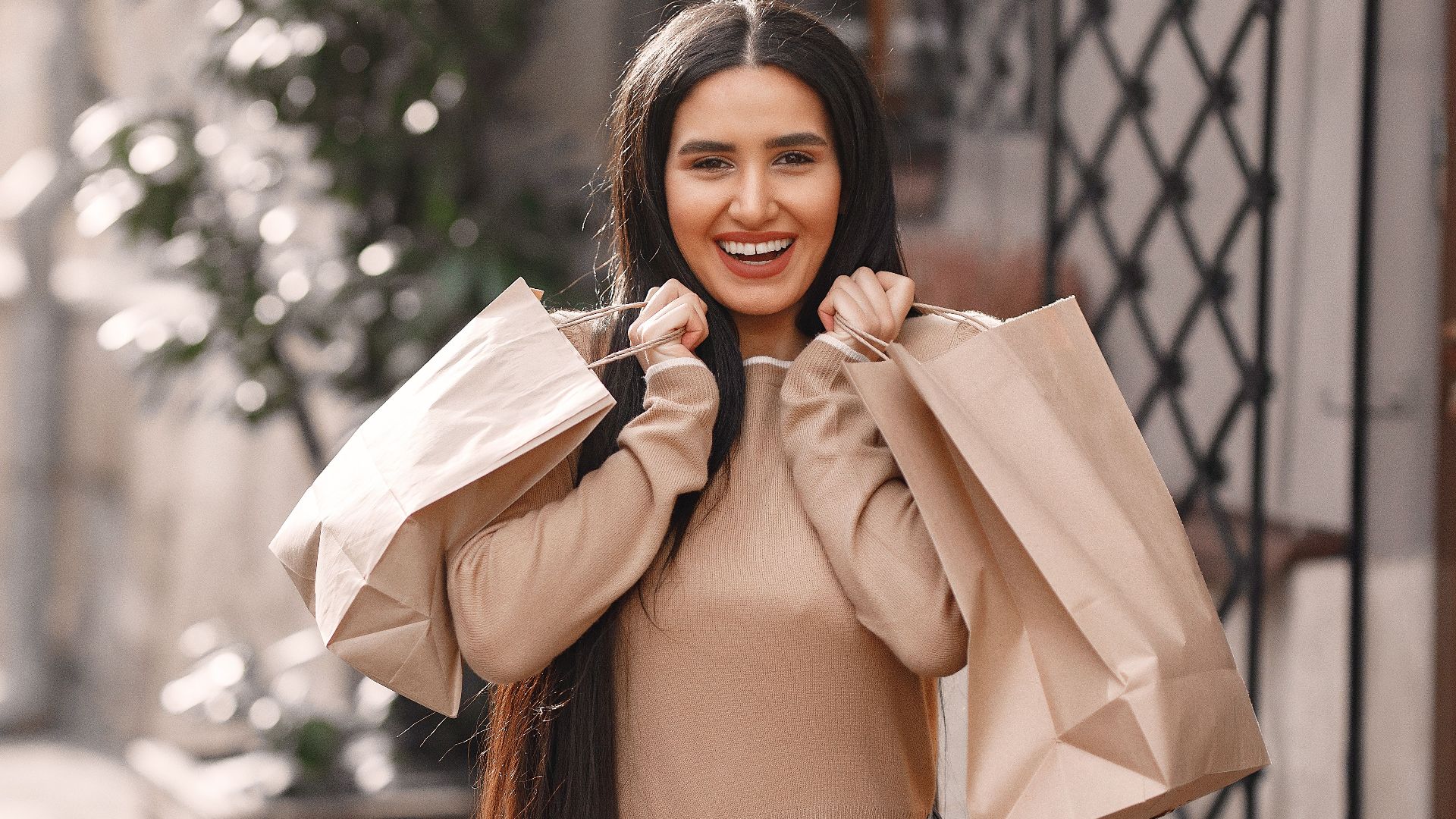 Young dark long haired woman in casual clothes holding paper bags and toothy smiling while looking at camera after shopping