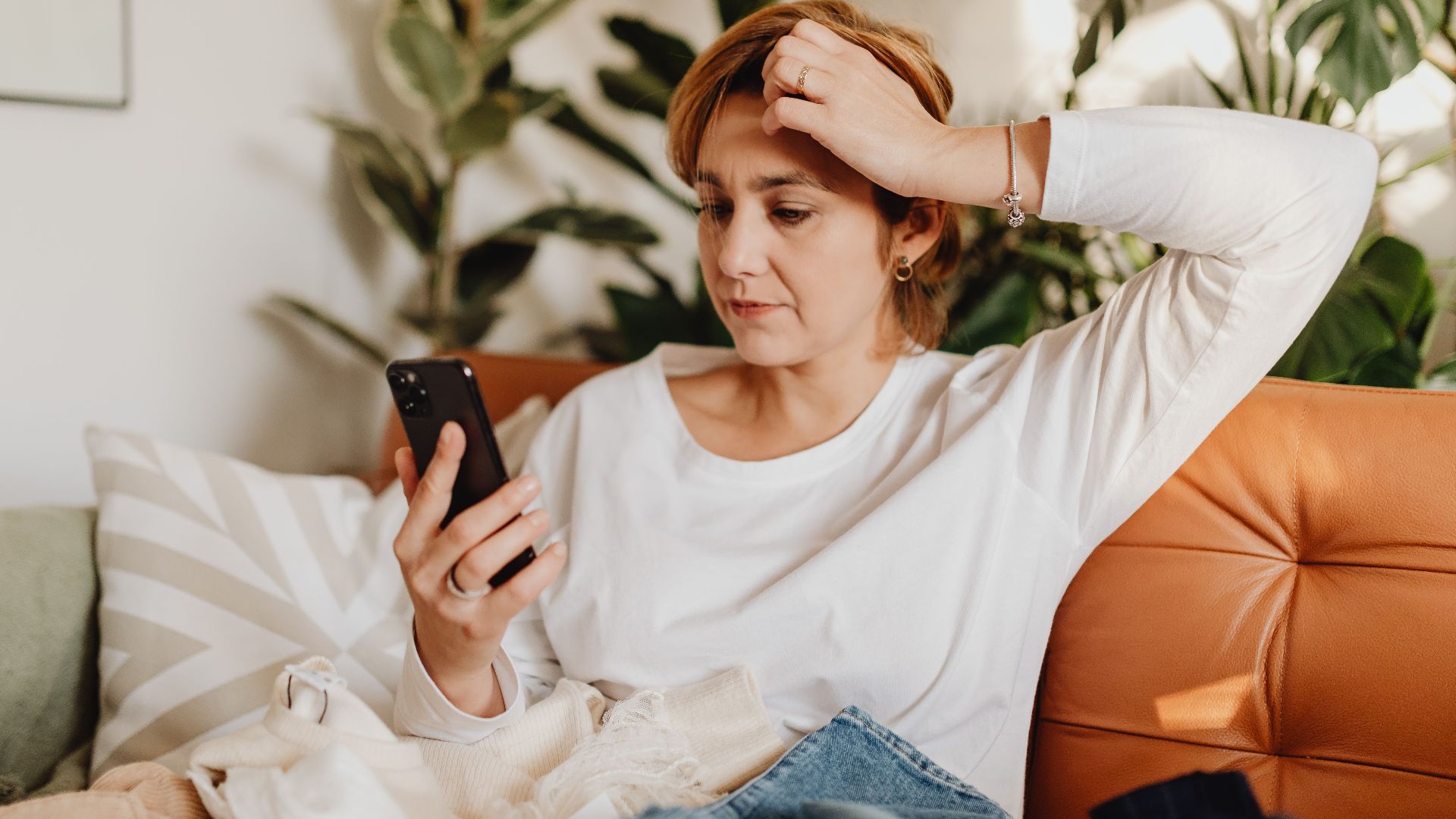 Young woman in white blouse using smartphone while sitting on an orange sofa surrounded by plants.
