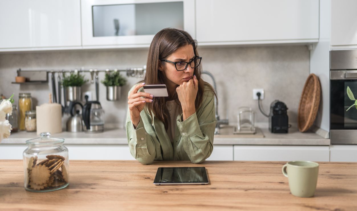 Shutterstock - 2696379511 - Anxious young woman deciding whether to pay online—holding a credit card and staring at a tablet concerned about fraud, hidden fees, and tight budgeting while shopping from home.