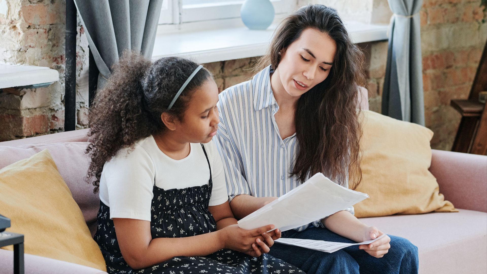 A woman and young girl sitting on a couch reading documents in a cozy living room.