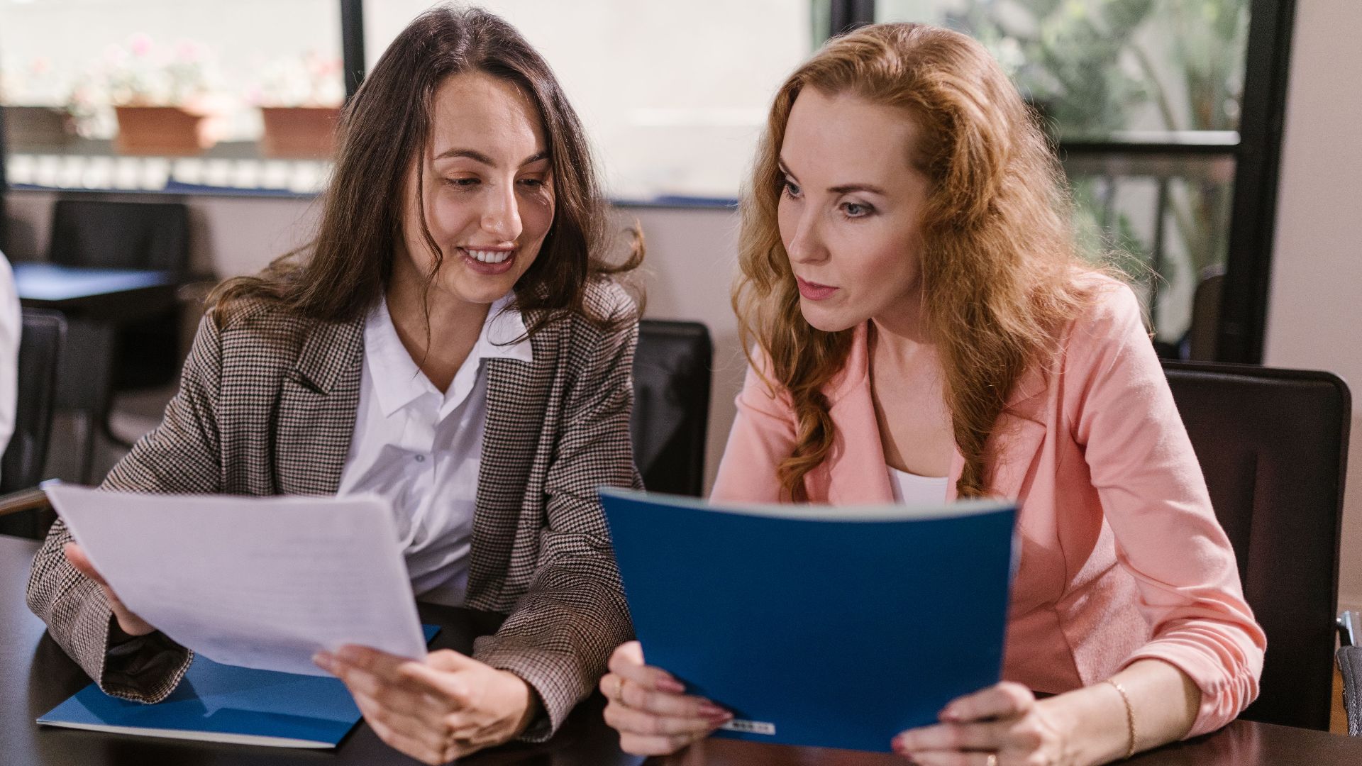 Two businesswomen reviewing documents together in an office setting, emphasizing teamwork and cooperation.