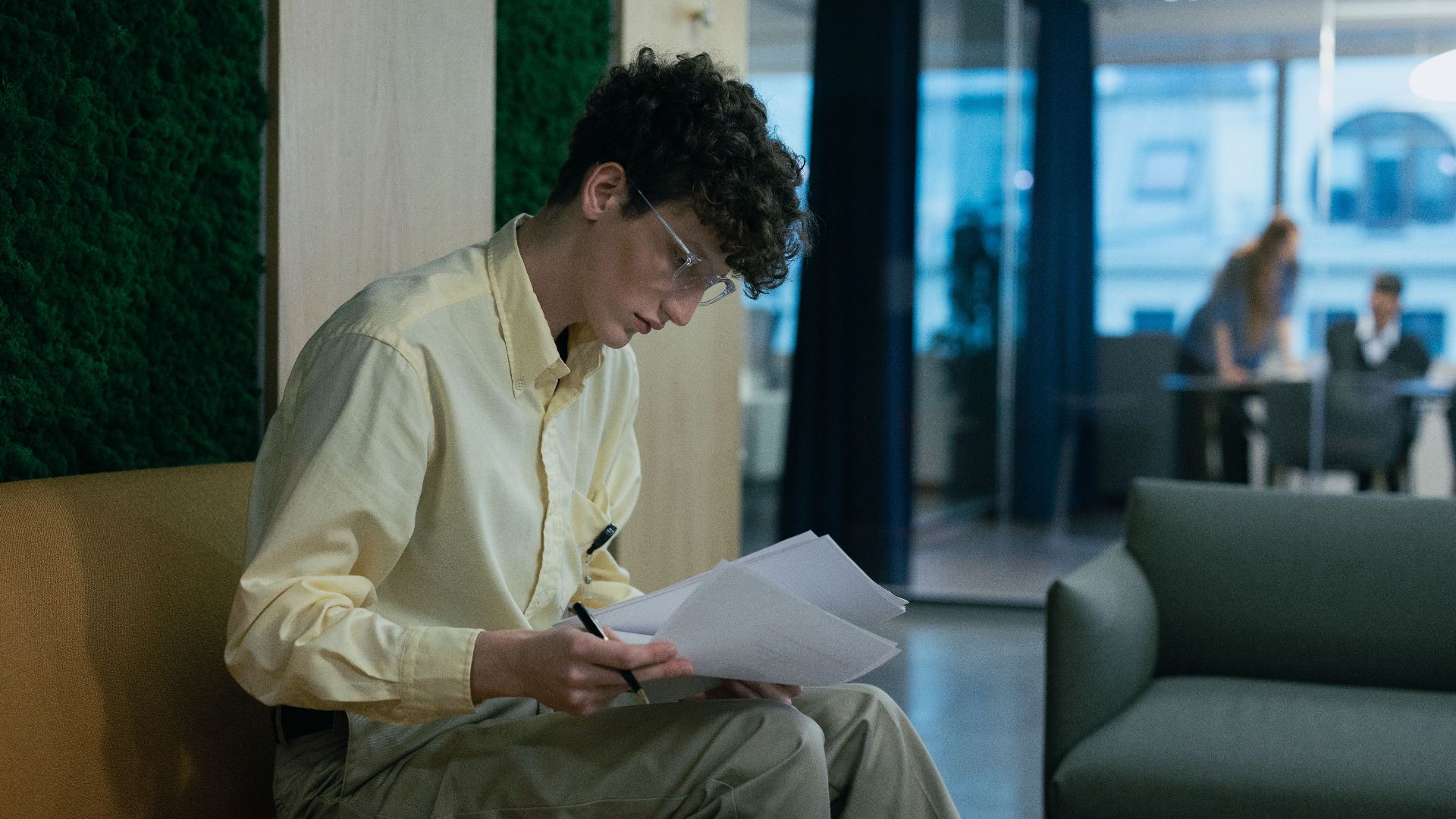 A focused young professional reading documents on a couch in a modern office environment.