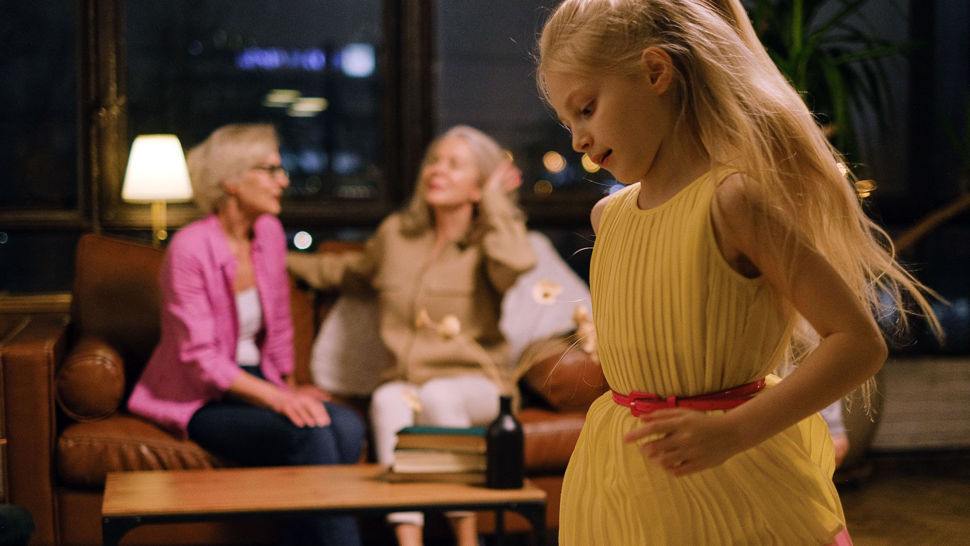 A joyful young girl in a yellow dress dances inside while family members relax on the couch.