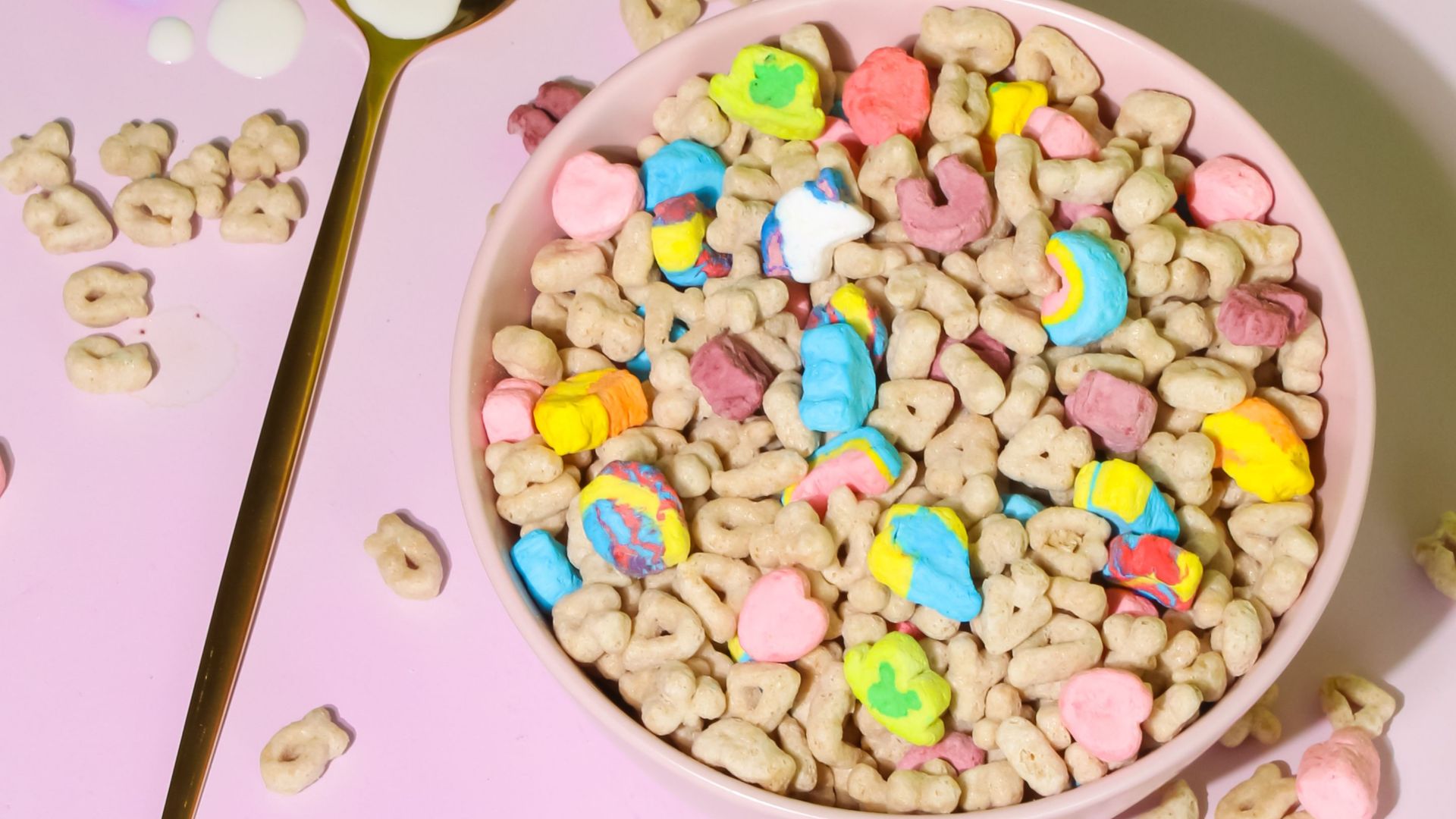 Bright and playful breakfast scene of colorful cereal in a pink bowl with a gold spoon on a pastel backdrop.