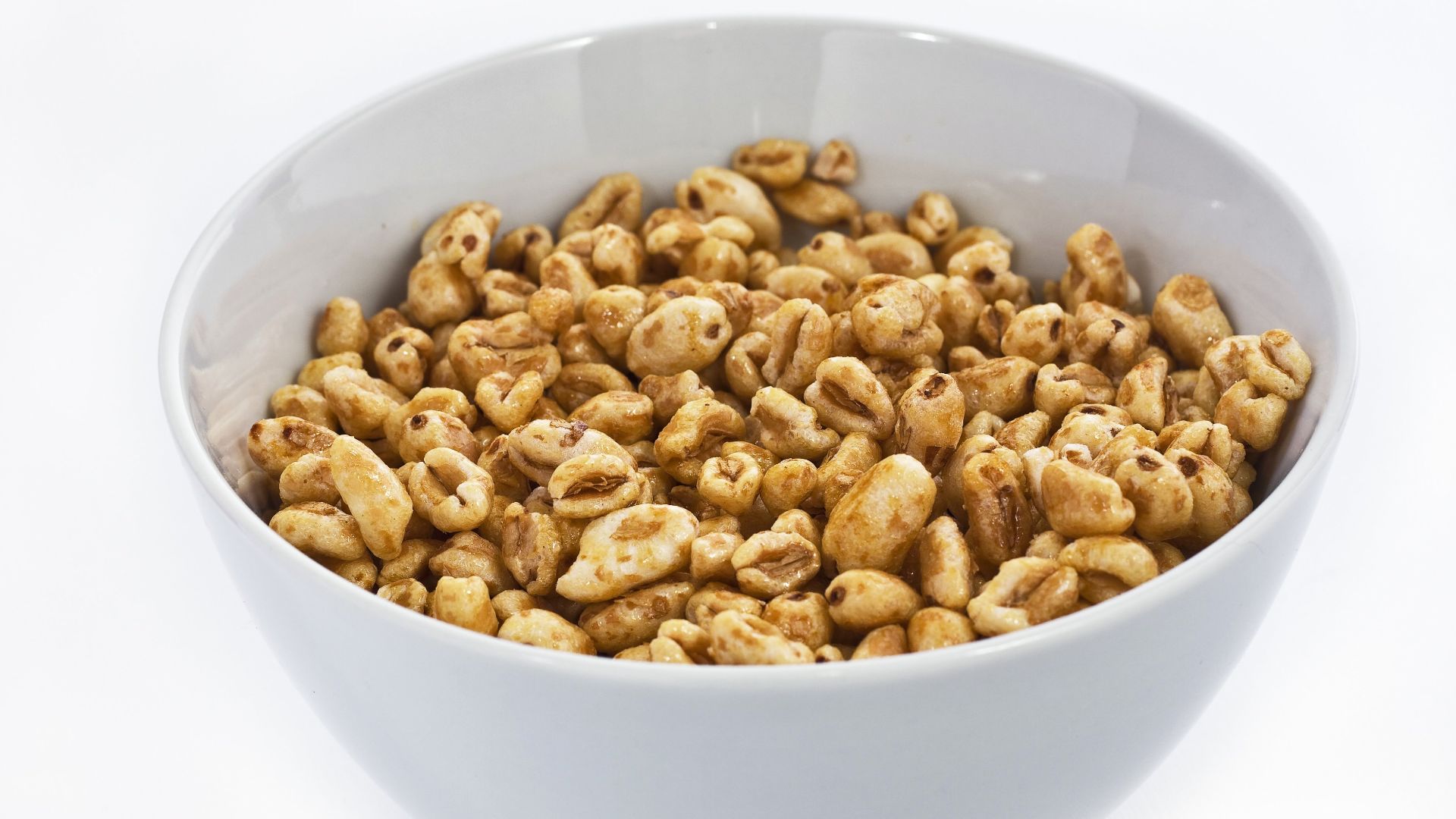 Simple, one color small sized ceramic bowl filled with many crunchy honey cereals, on a white background. The cereals have a various peanut shapes and sizes and light brown tones.