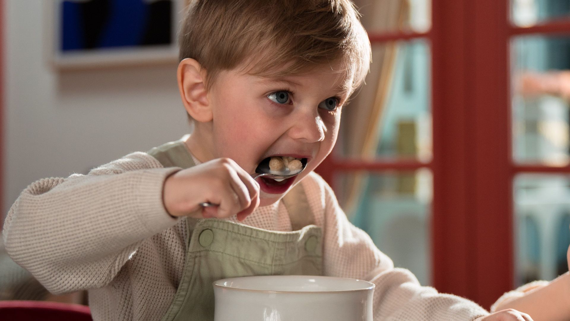 A young boy eating breakfast cereal with enthusiasm at home, seated at a kitchen table indoors.
