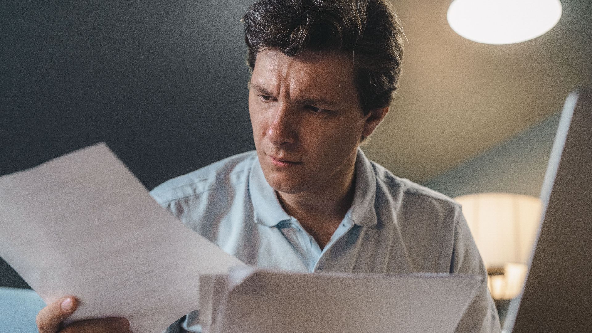 A man intently examines papers, seated indoors under warm lighting, focusing on his work.