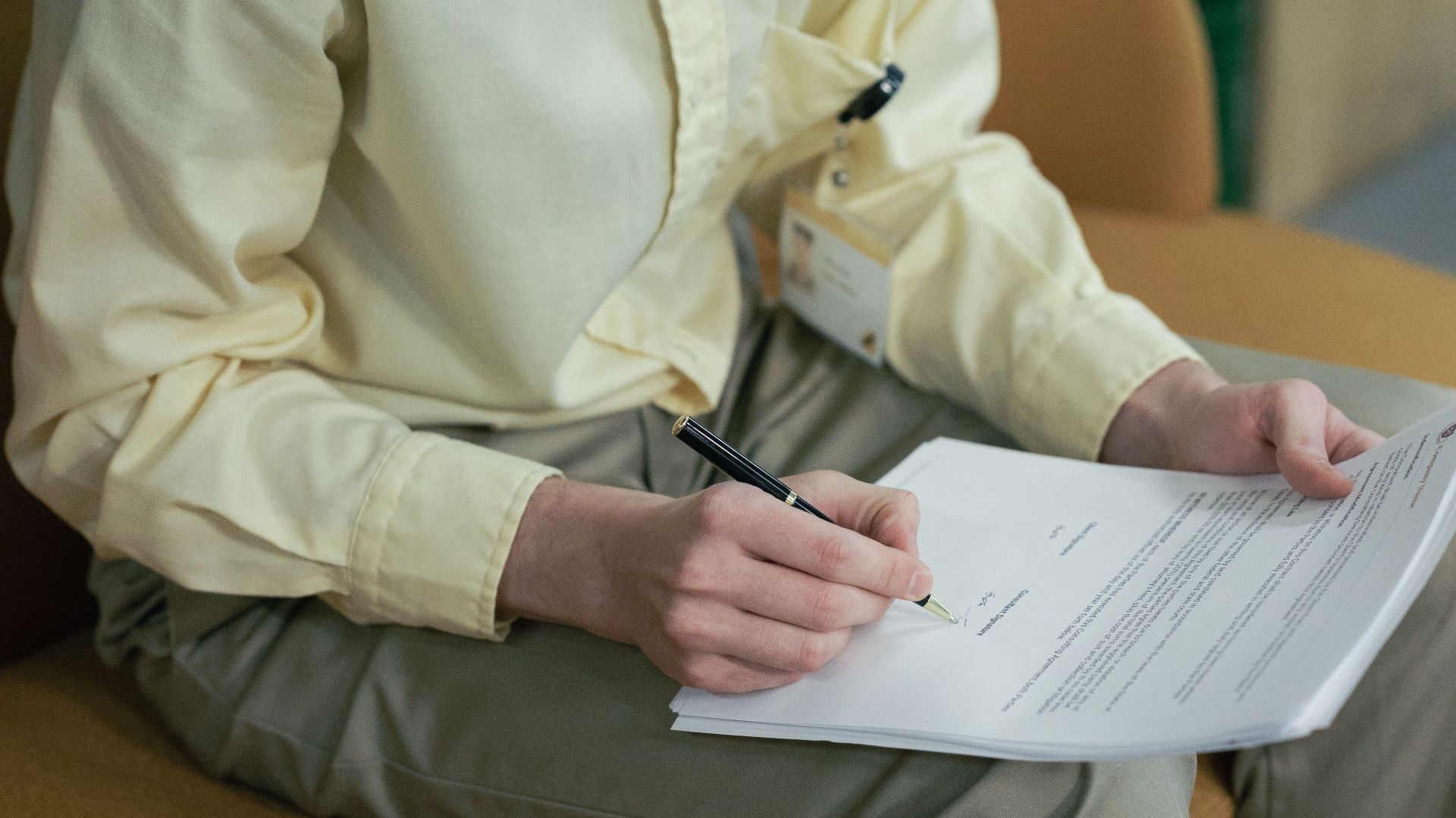A young man with curly hair and glasses signs papers while sitting indoors.