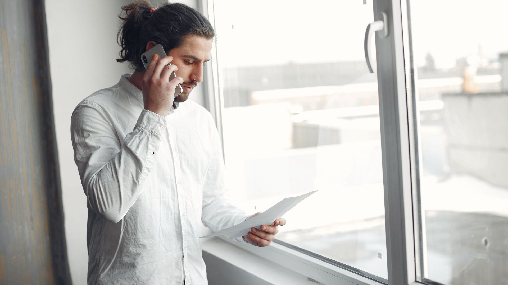 Young man in white shirt, on phone call holding a document, standing by a large window.