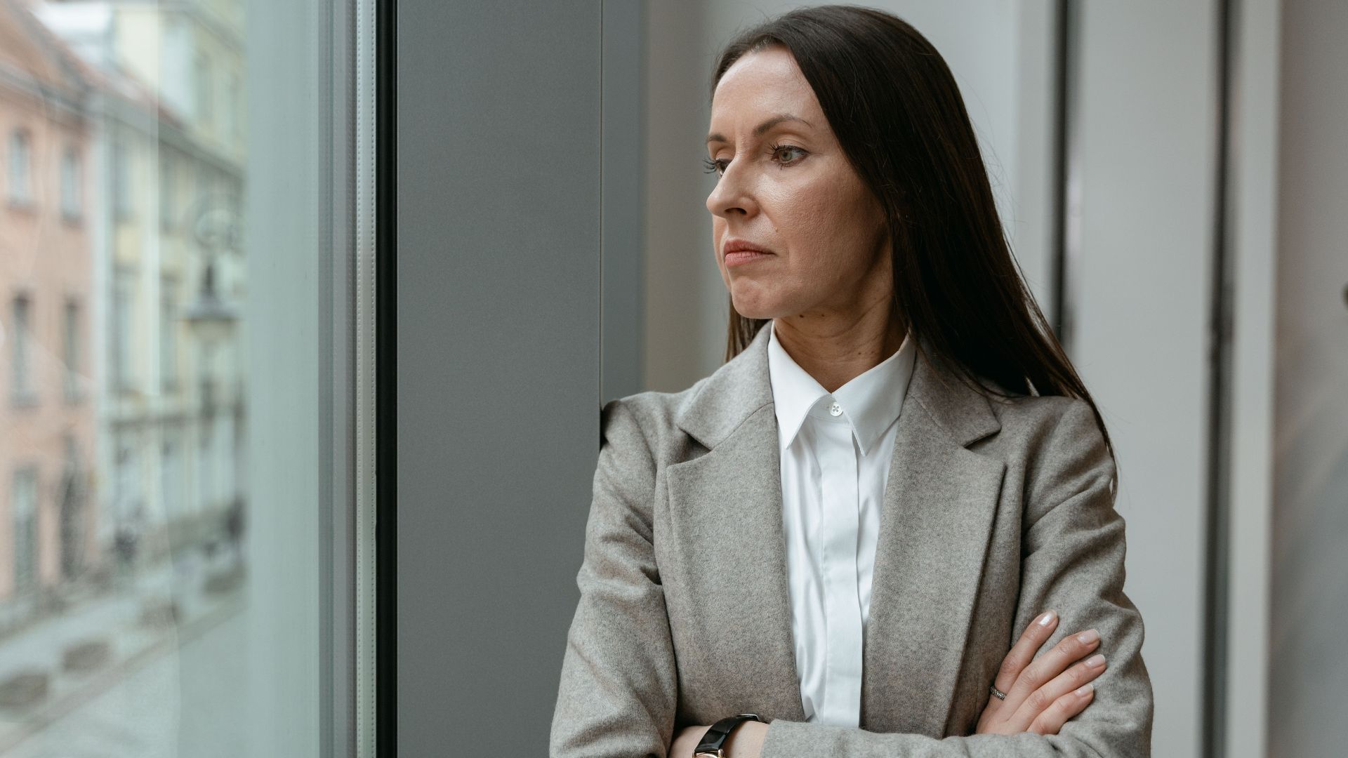 A professional woman in a gray blazer stands with arms crossed, gazing out an office window.