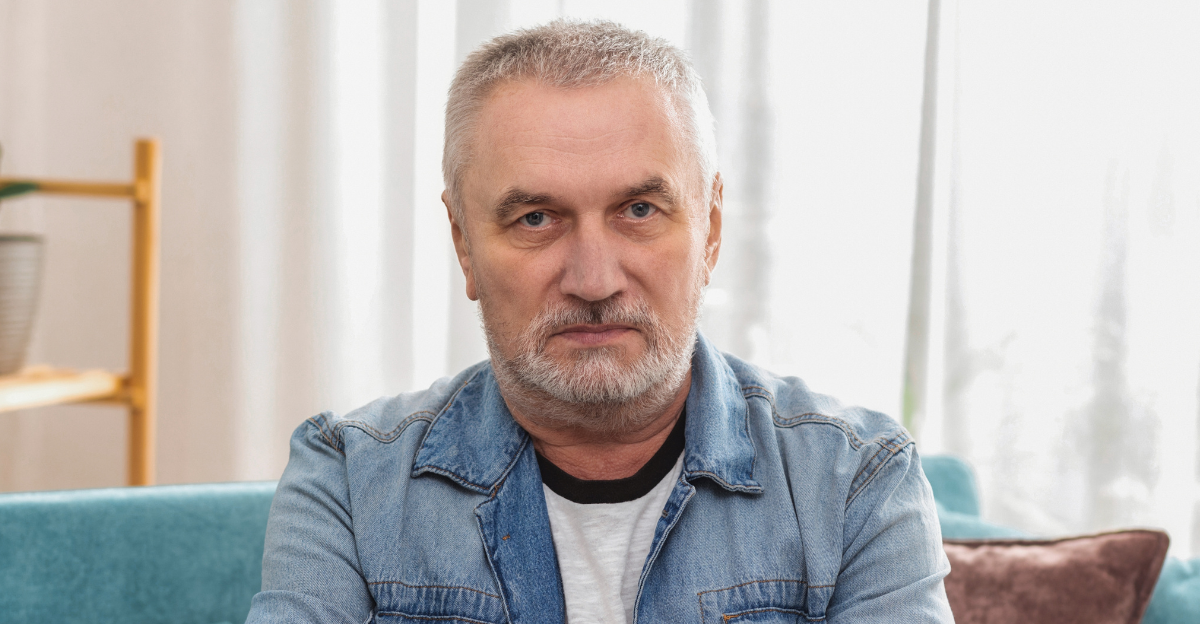 Angry senior man with gray hair and crossed arms looking at camera, sitting on sofa at home 