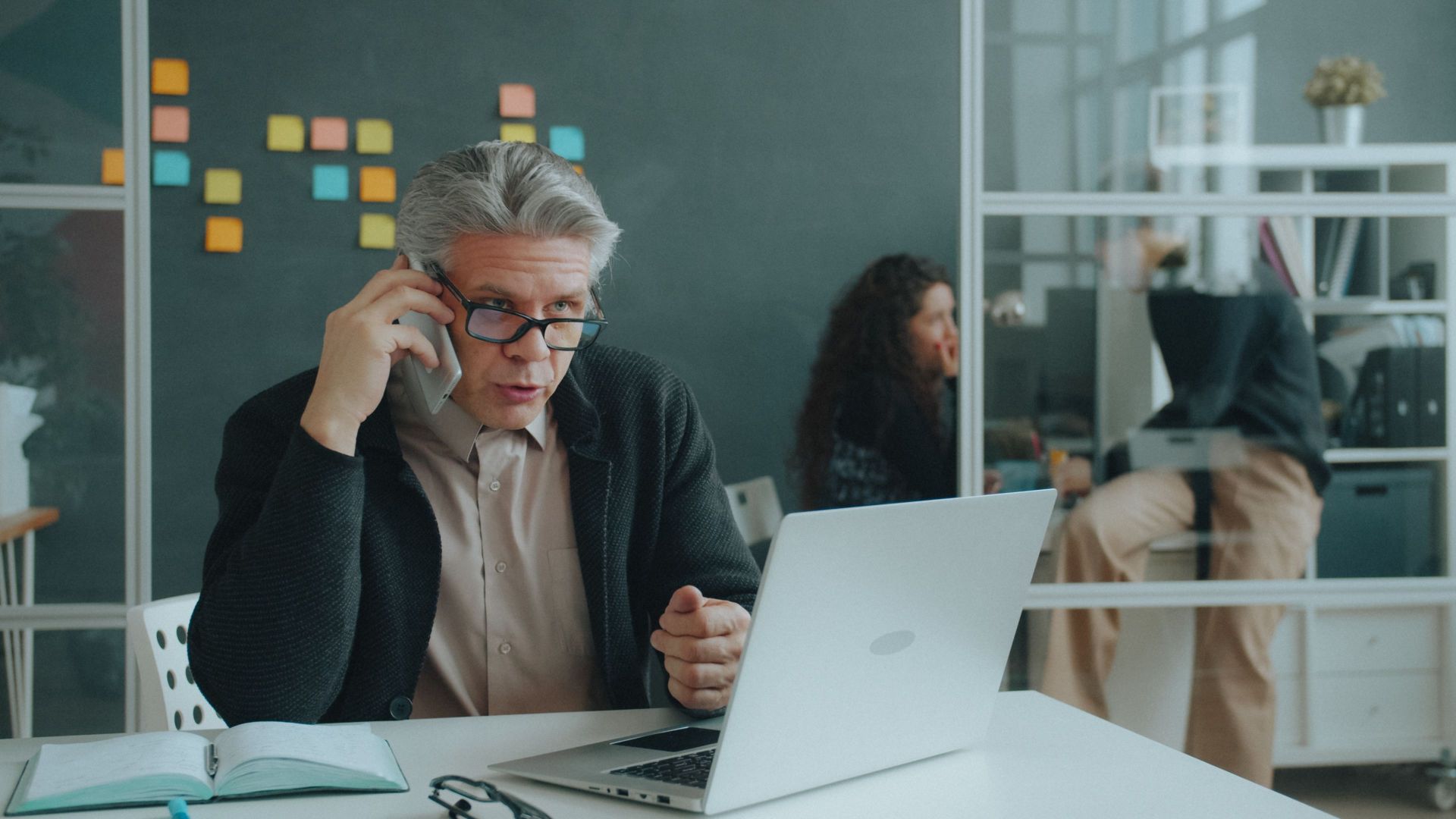Senior man discussing business on phone with laptop in modern office.