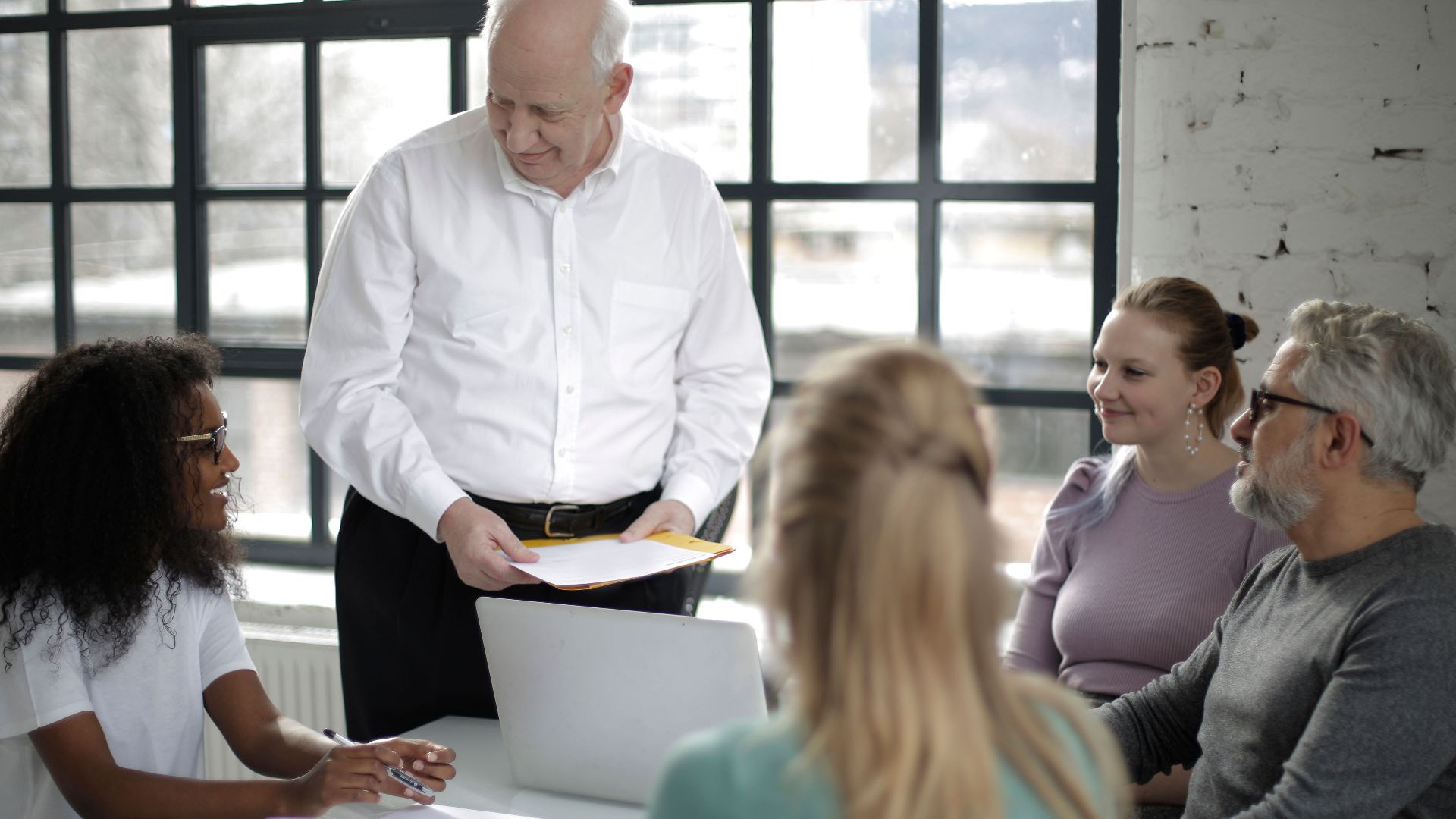 Positive multiracial colleagues of different ages gathering at table in modern workspace while old man presentation project
