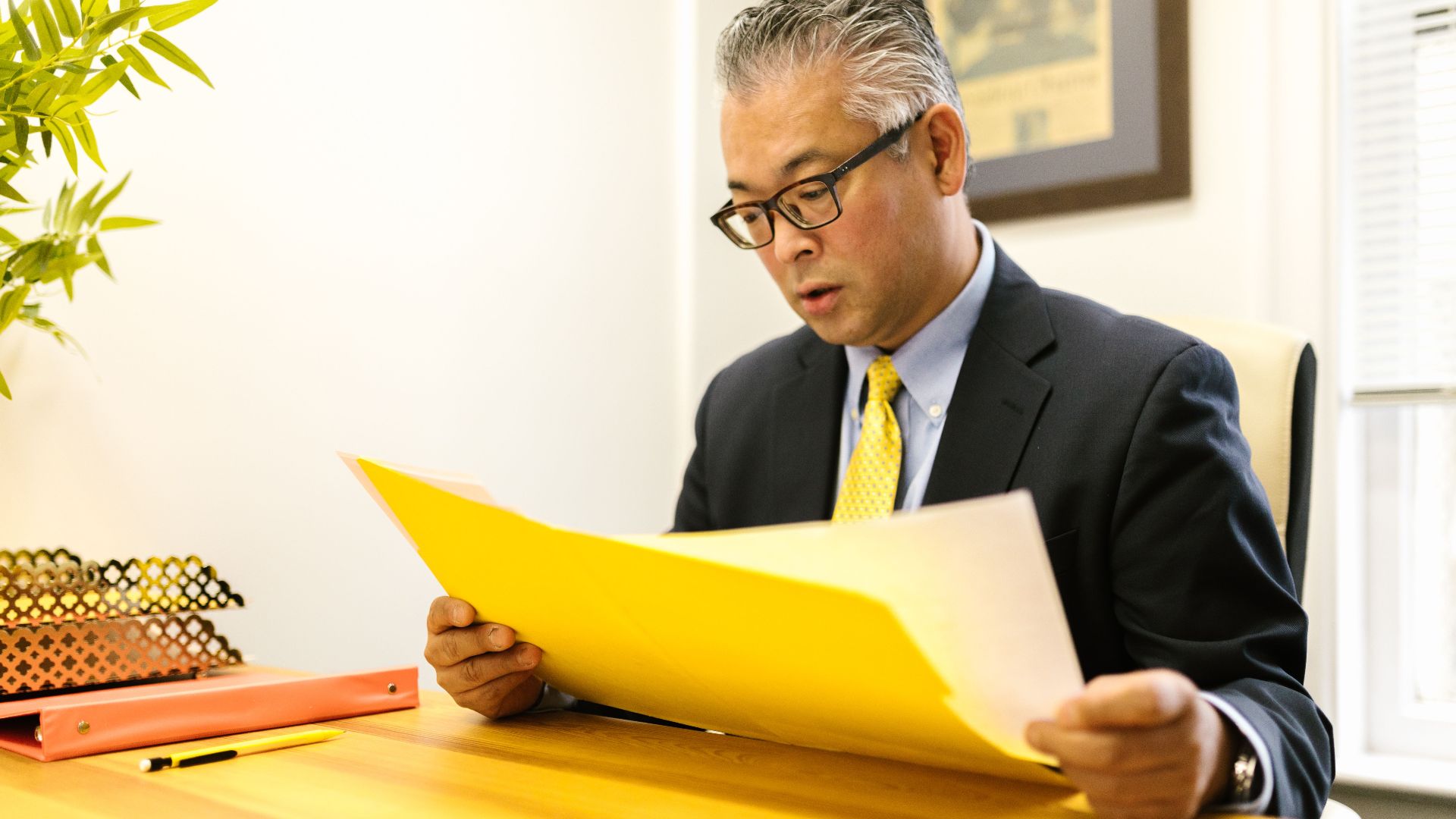 Asian businessman in corporate attire reading documents at office desk with a yellow folder.