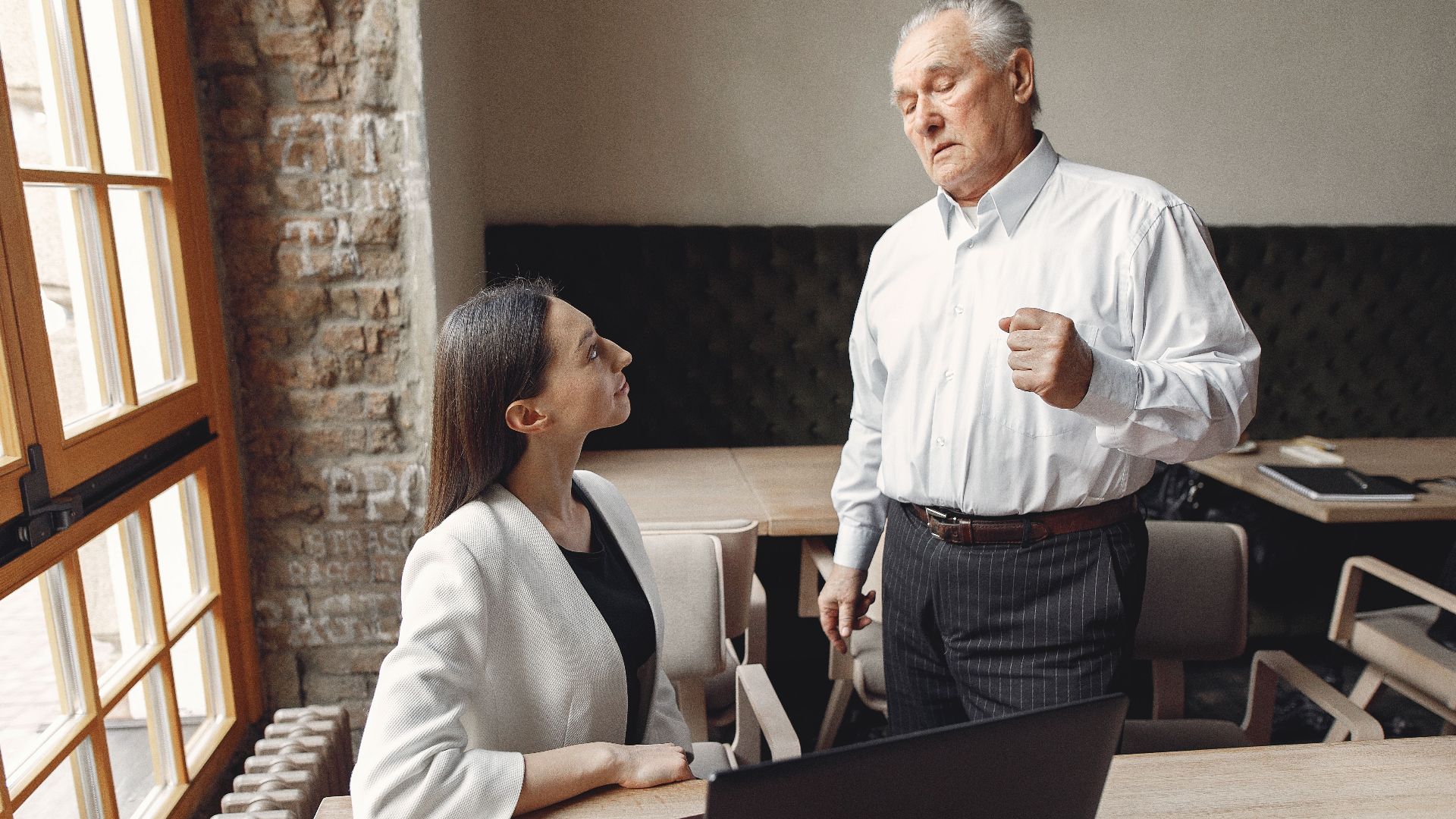 Aged businessman in formal wear talking with well dressed female employee with laptop and cup of coffee about business issue in modern office