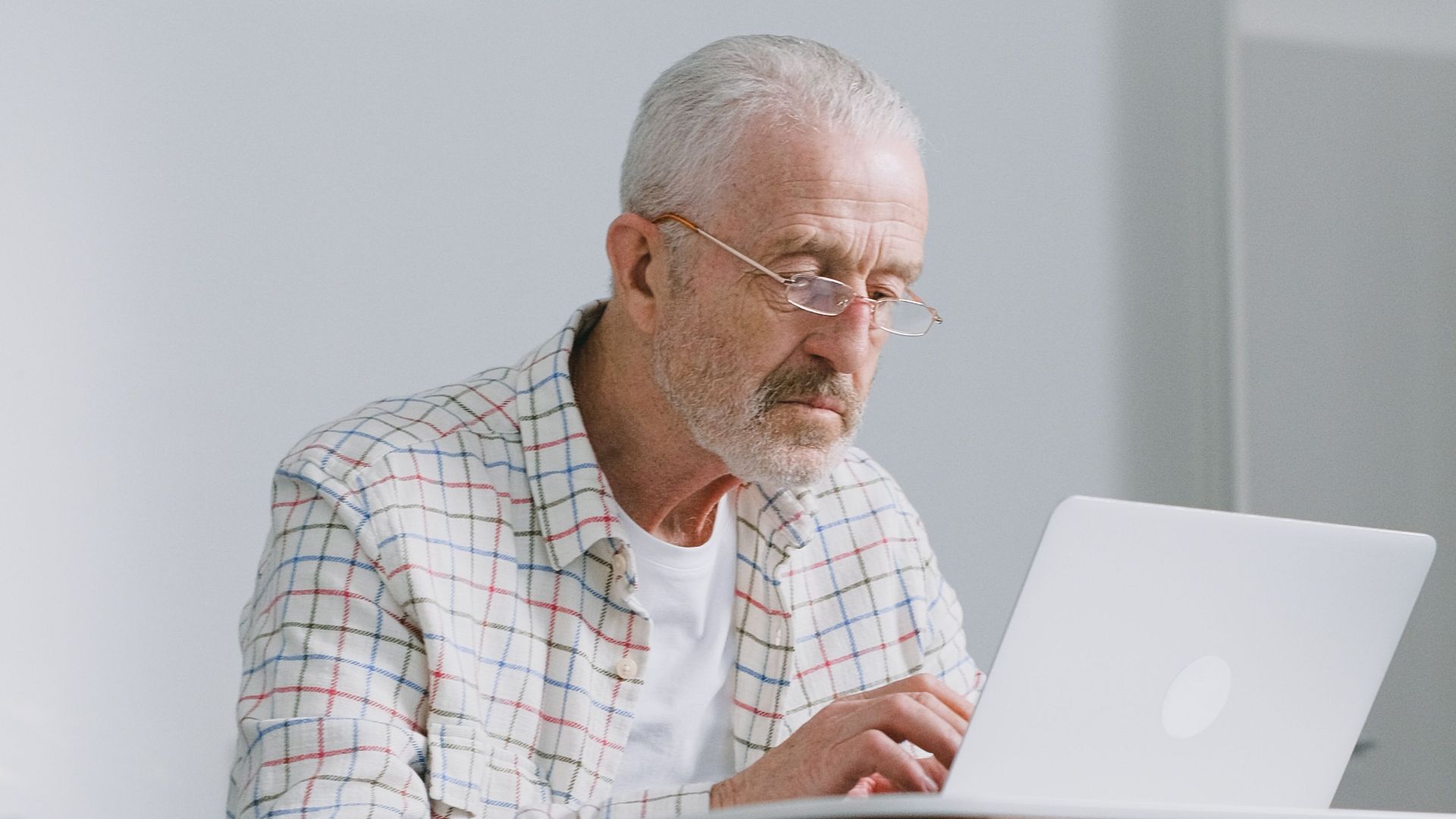 Elderly man focused on typing on laptop indoors. Natural lighting, modern ambiance.