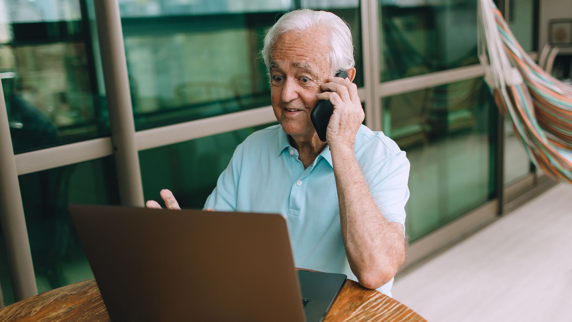 Elderly man multitasking on laptop and phone indoors, focused on technology.
