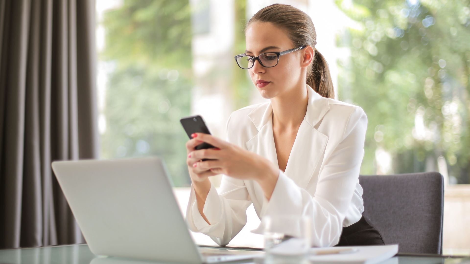 Concentrated young woman in formal clothes and eyeglasses sitting at glass table with laptop while surfing smartphone