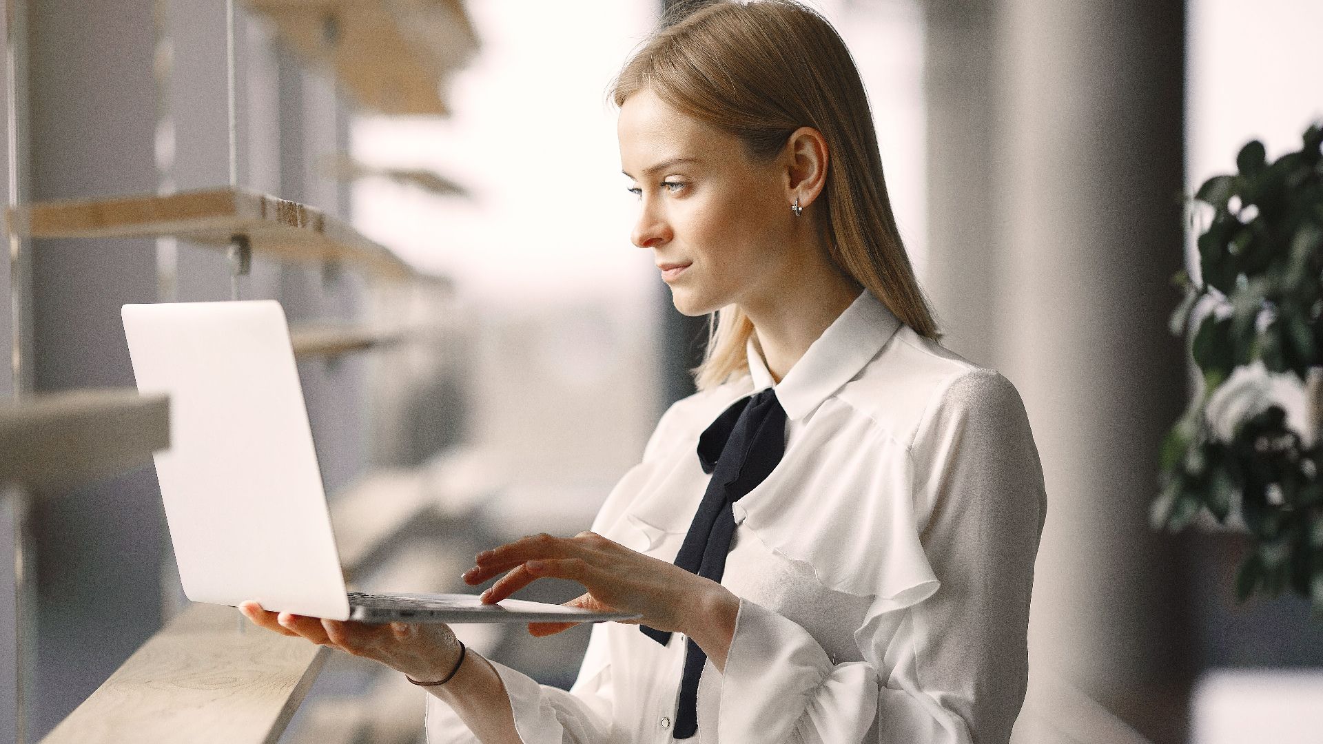 Focused female employee in formal clothes using laptop near window decoration while standing in office lobby