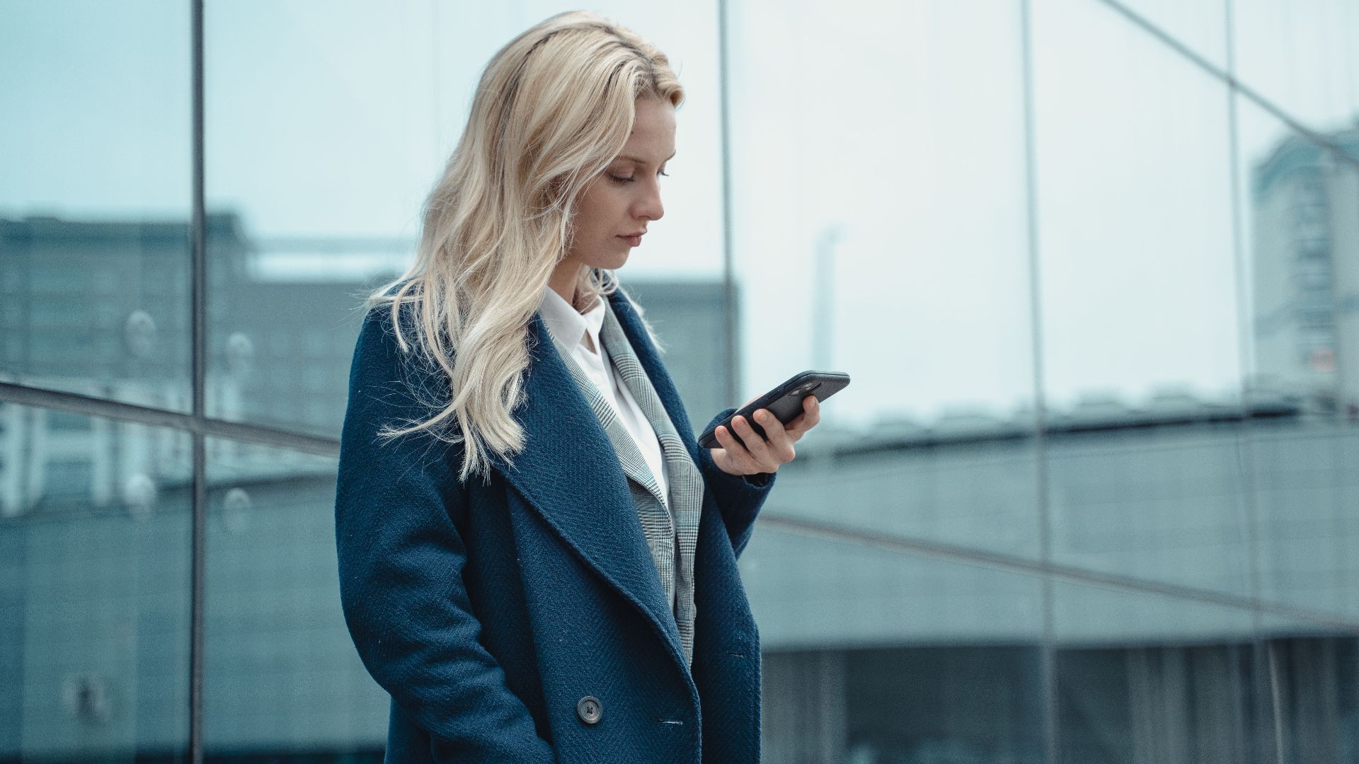 Professional woman in blue coat using smartphone outside office building.