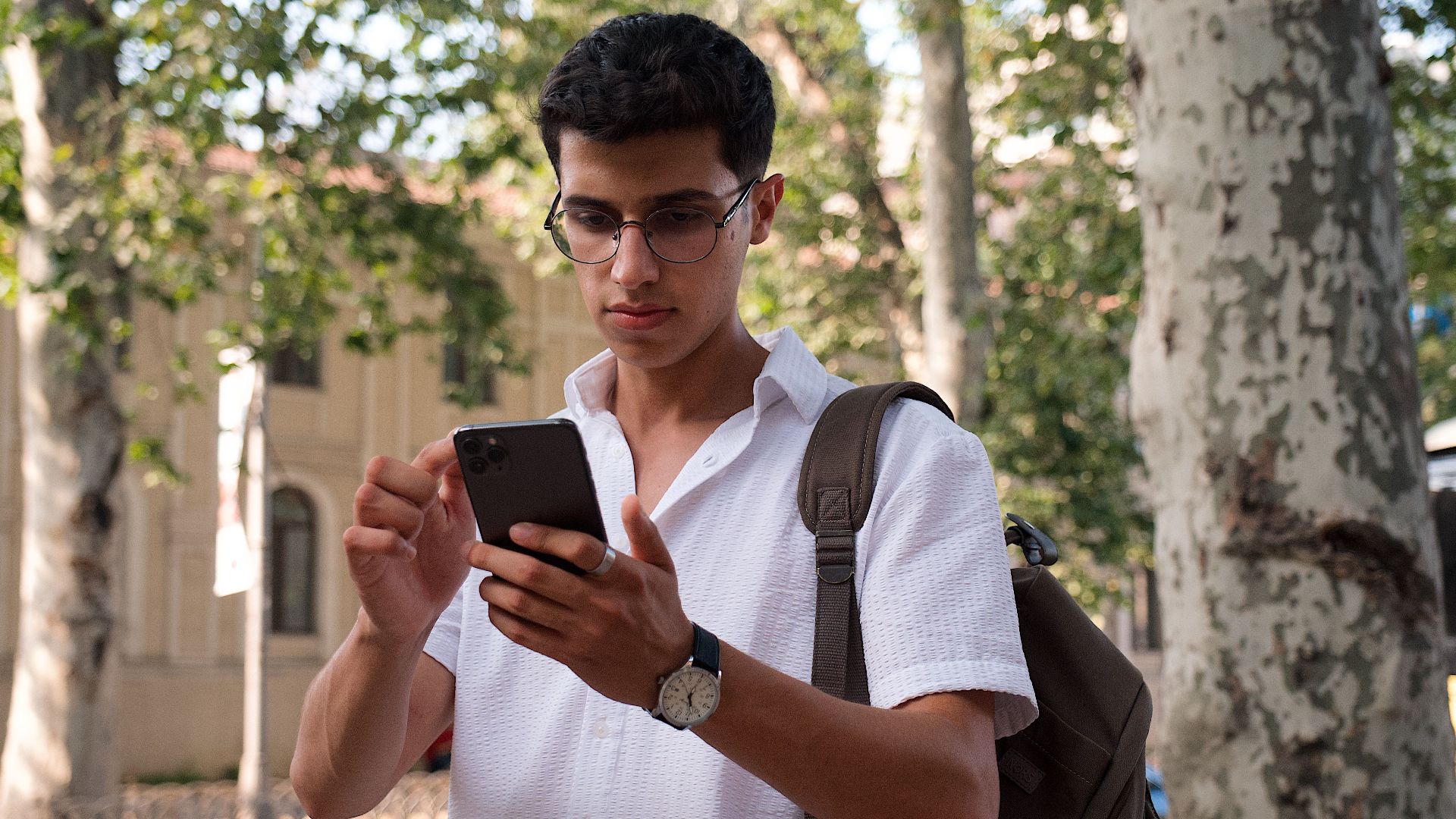 Young man using smartphone on a city street, wearing eyeglasses and a backpack.