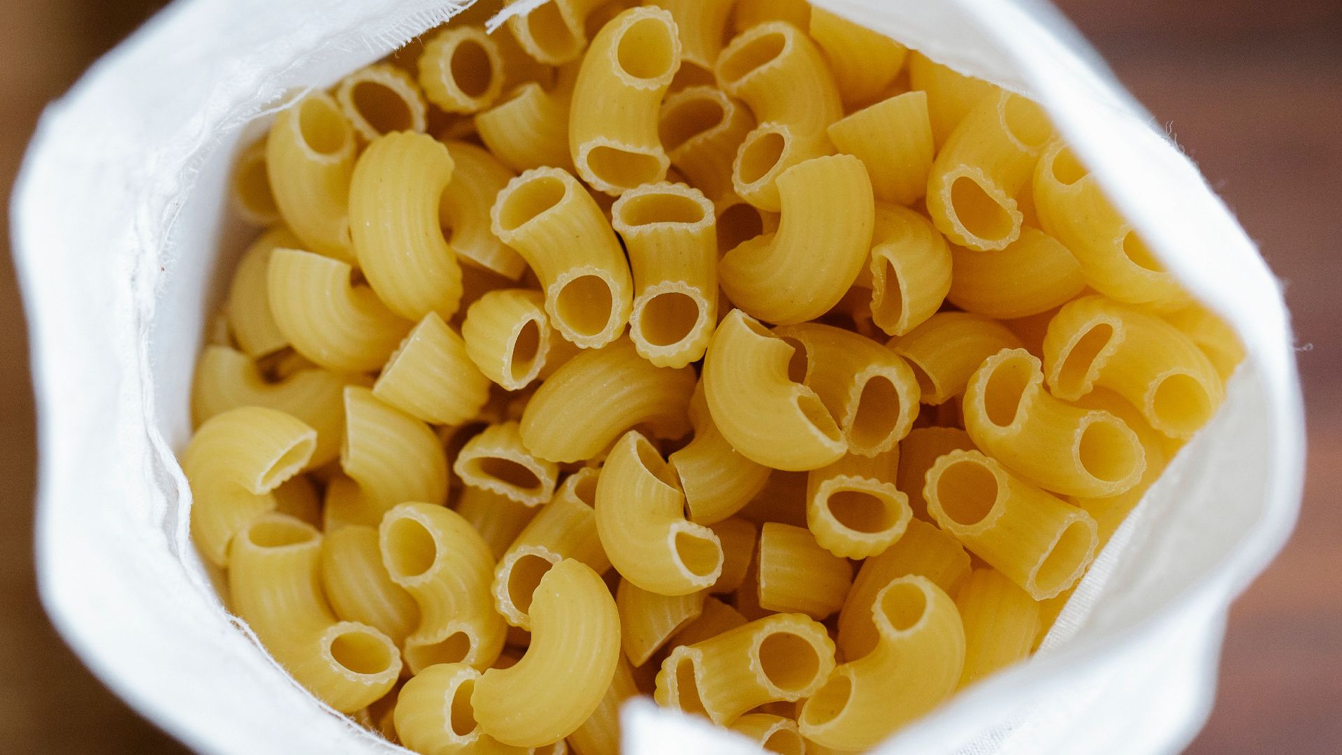Top view white eco friendly sack full of uncooked macaroni placed on table on blurred background in light kitchen at home