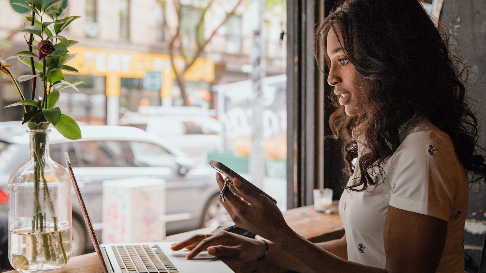 Afro American woman working remotely on laptop and phone in urban café setting.