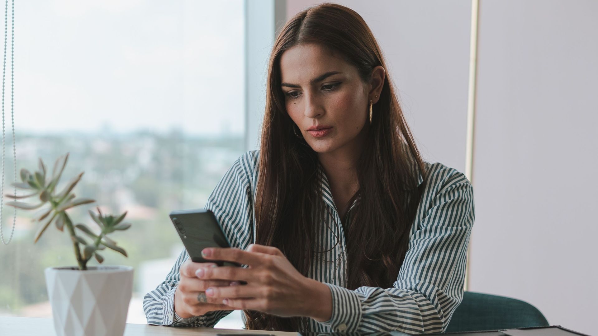 A businesswoman attentively using her smartphone at a desk in a modern office setting with a potted plant.