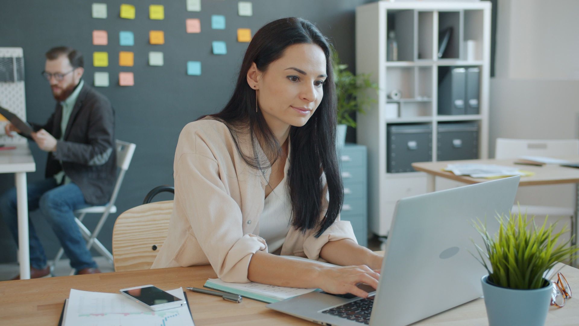 Woman working on laptop in a modern office with a colleague in the background.