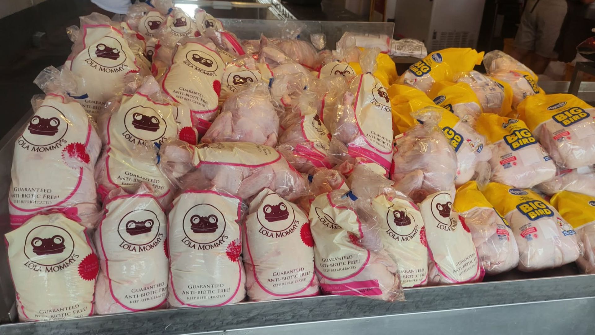 This indoor eye-level photograph captures a large, organized display of packaged whole chickens resting on a metallic counter at a local market stall. The left side features white plastic bags branded with the