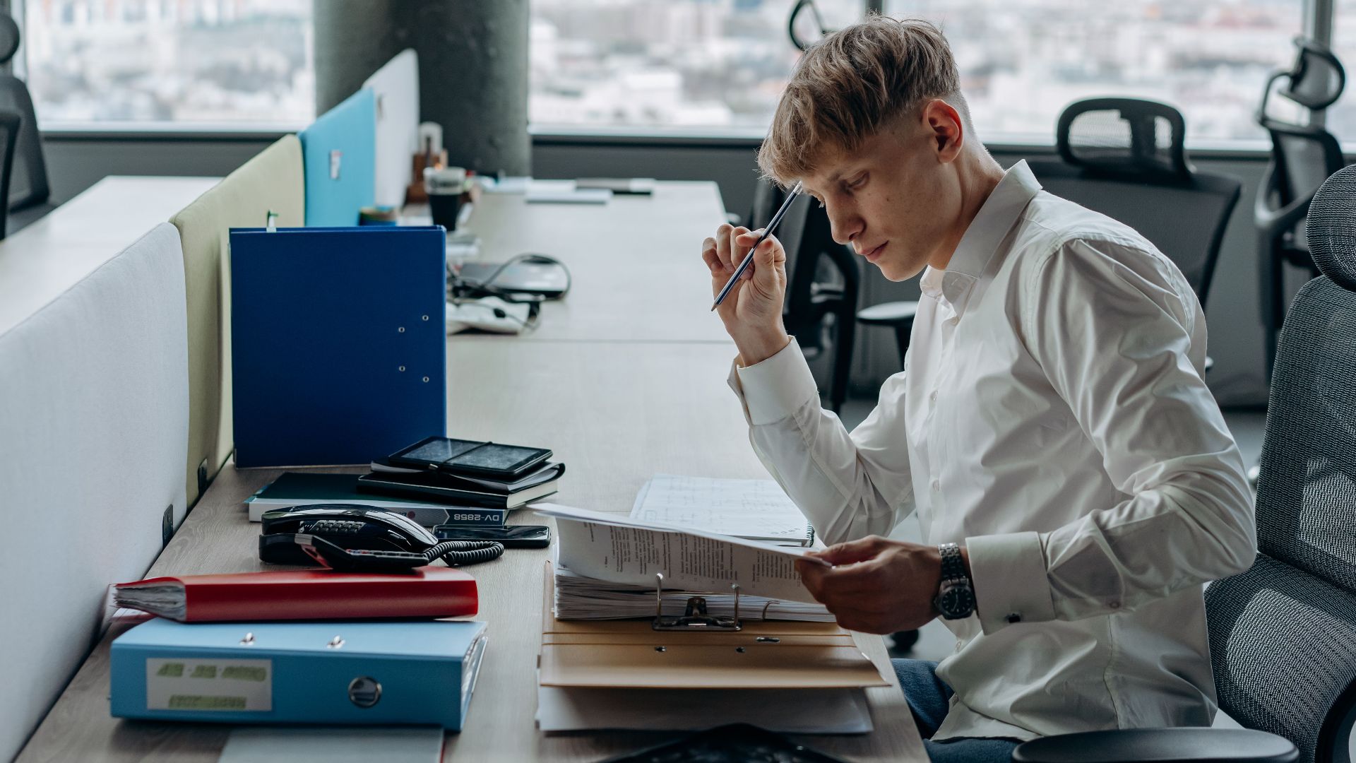 Man in white shirt concentrating on paperwork at modern office desk with city view.