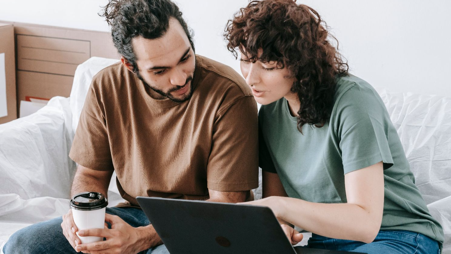 Young couple sits on a sofa using a laptop during a move, surrounded by boxes and sipping coffee.
