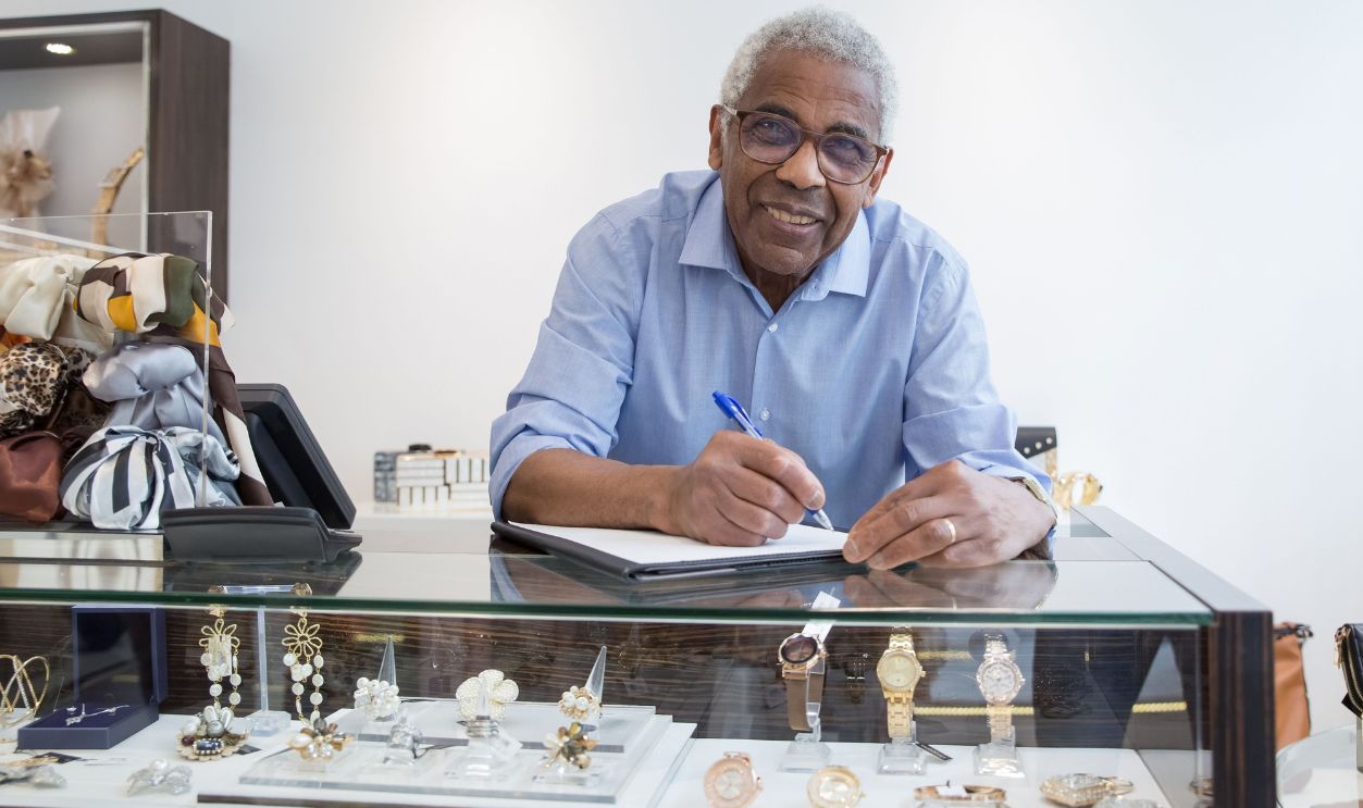 Smiling elderly shopkeeper in Portugal writing at jewelry display counter