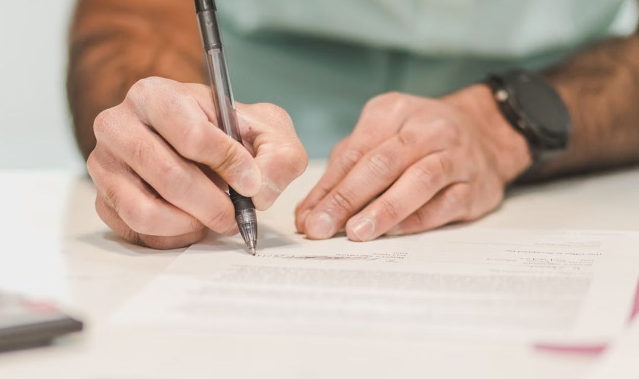 Close-up of a man signing an official document with a ballpoint pen on a desk.