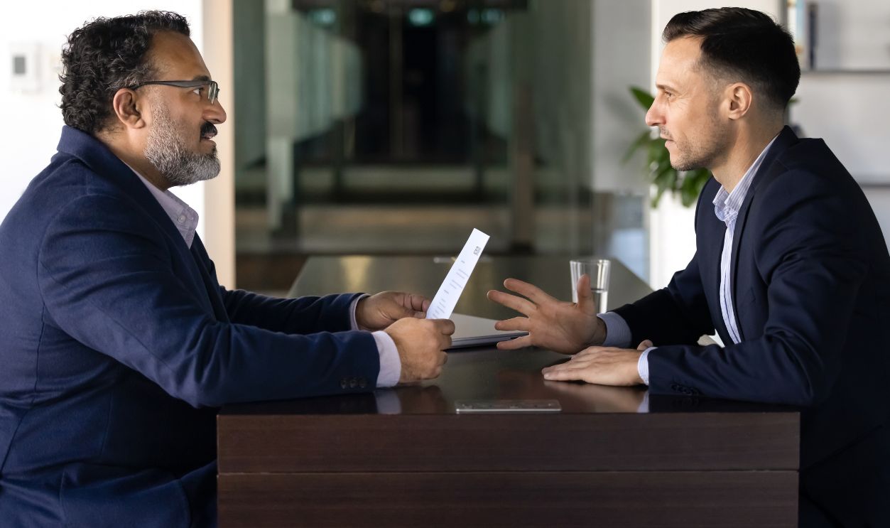 Two diverse male younger and older business coworkers arguing at work table, sitting opposite, talking, having conflict of generations. Senior boss dissatisfied with young professional paper report