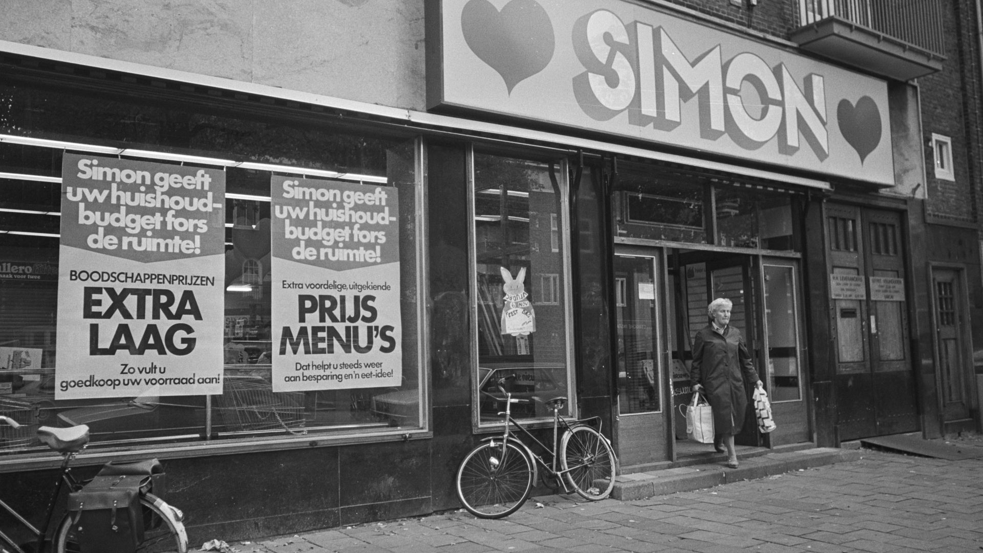 Black-and-white photo from 1981 of a woman walking out of the Simon de Wit supermarket in de Rijnstraat in Amsterdam with two bags full of groceries. Large advertisements are hanging in the shop windows. This is Simon de Wit's final years; the last store
