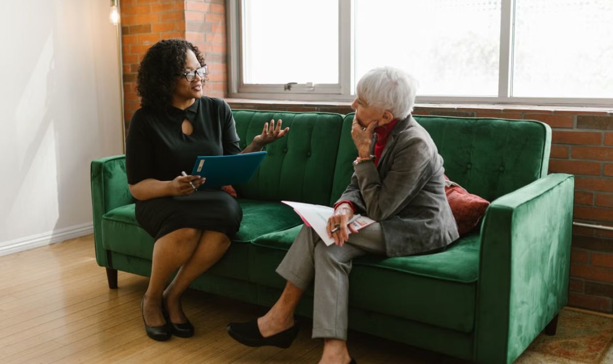 Two women engaged in a professional conversation sitting on a green sofa in a modern office setting.