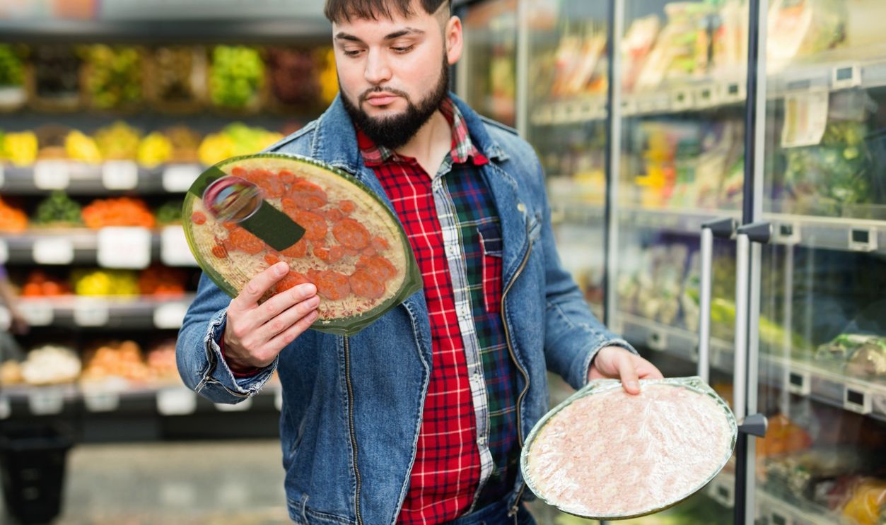 Young cheerful smiling man shopping at supermarket food department, choosing frozen pizzas