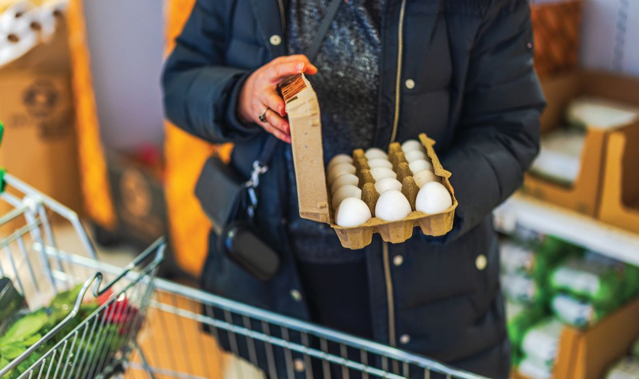Woman in black winter coat holding carton of white eggs while shopping in grocery store aisle. Sweden.