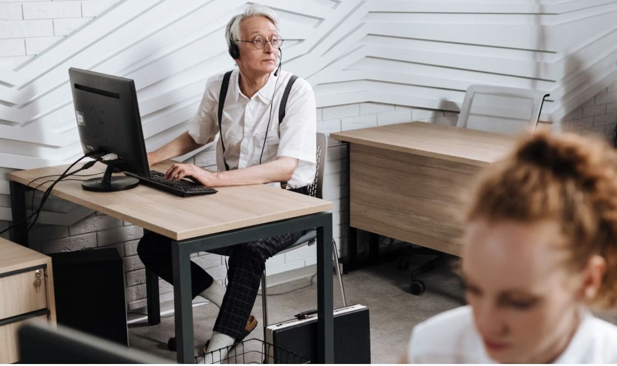 Senior call center agent using a headset and computer in a modern office setting.
