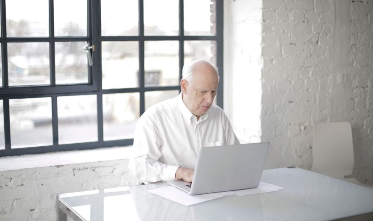 Elderly man working in a modern white office with laptop. Professional and focused environment.