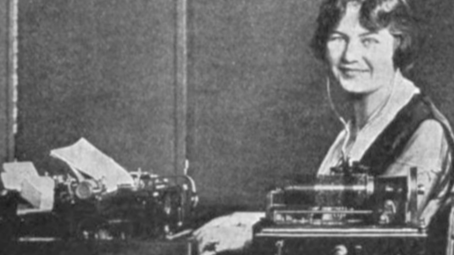 Transcribing dictation from a Dictaphone wax cylinder dictation machine, from the 1920s.  The dictation machine is in the foreground.  The typist is wearing 'stethoscope' type earphones attached to the machine.  Her foot is on a pedal which starts and sto
