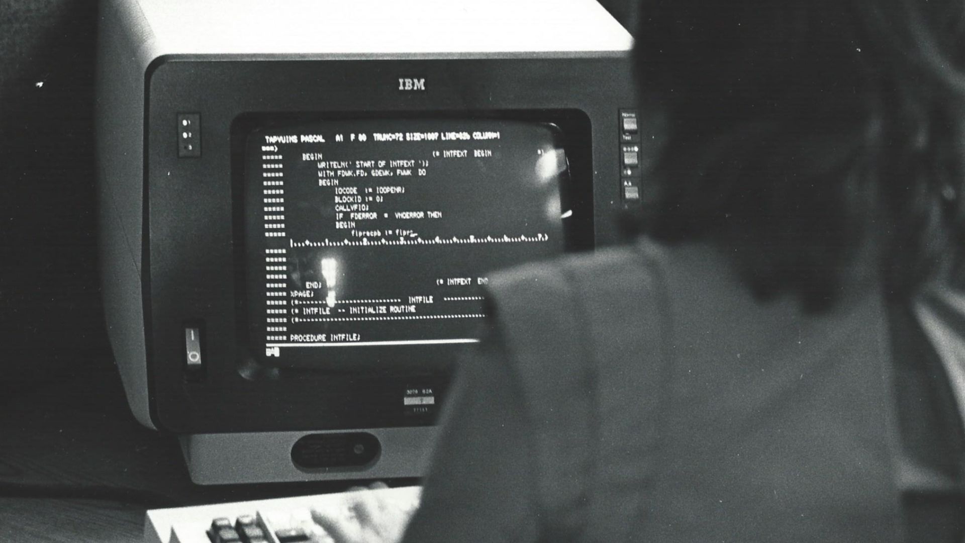 An Informatics General computer programmer working in a cubicle at the company's New York City office. She is looking at Pascal language code for the TAPS product on an IBM 3270 terminal.