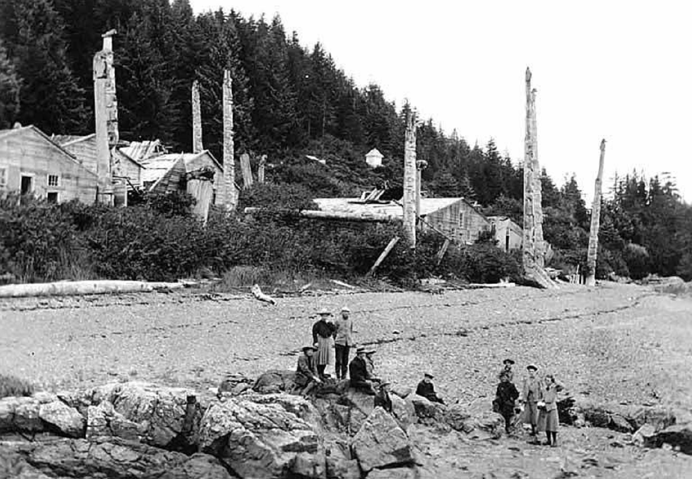 People standing on beach with Haida totem poles and dwellings in background - 1899