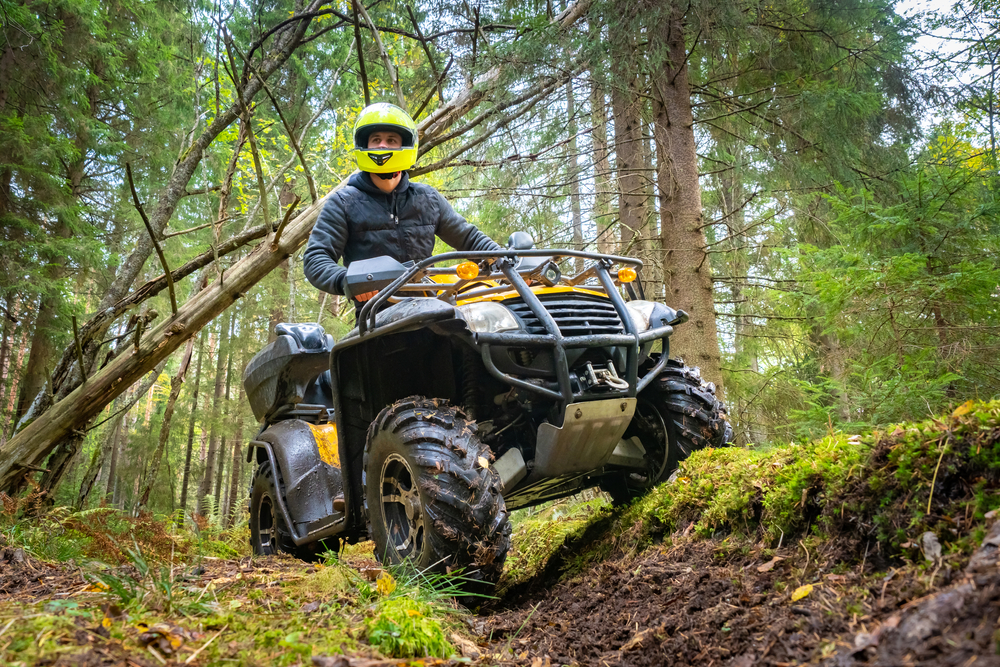 A man in a yellow helmet rides a Quad bike through the woods.