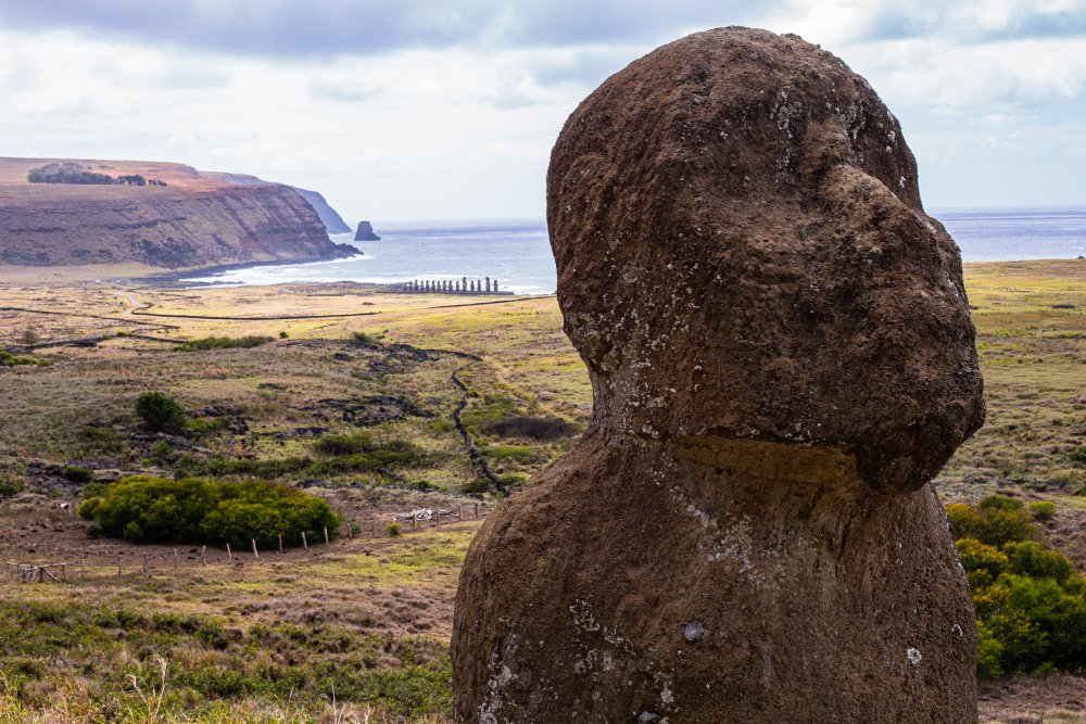 A large, weathered moai statue stands in the foreground, its gaze directed towards the ocean