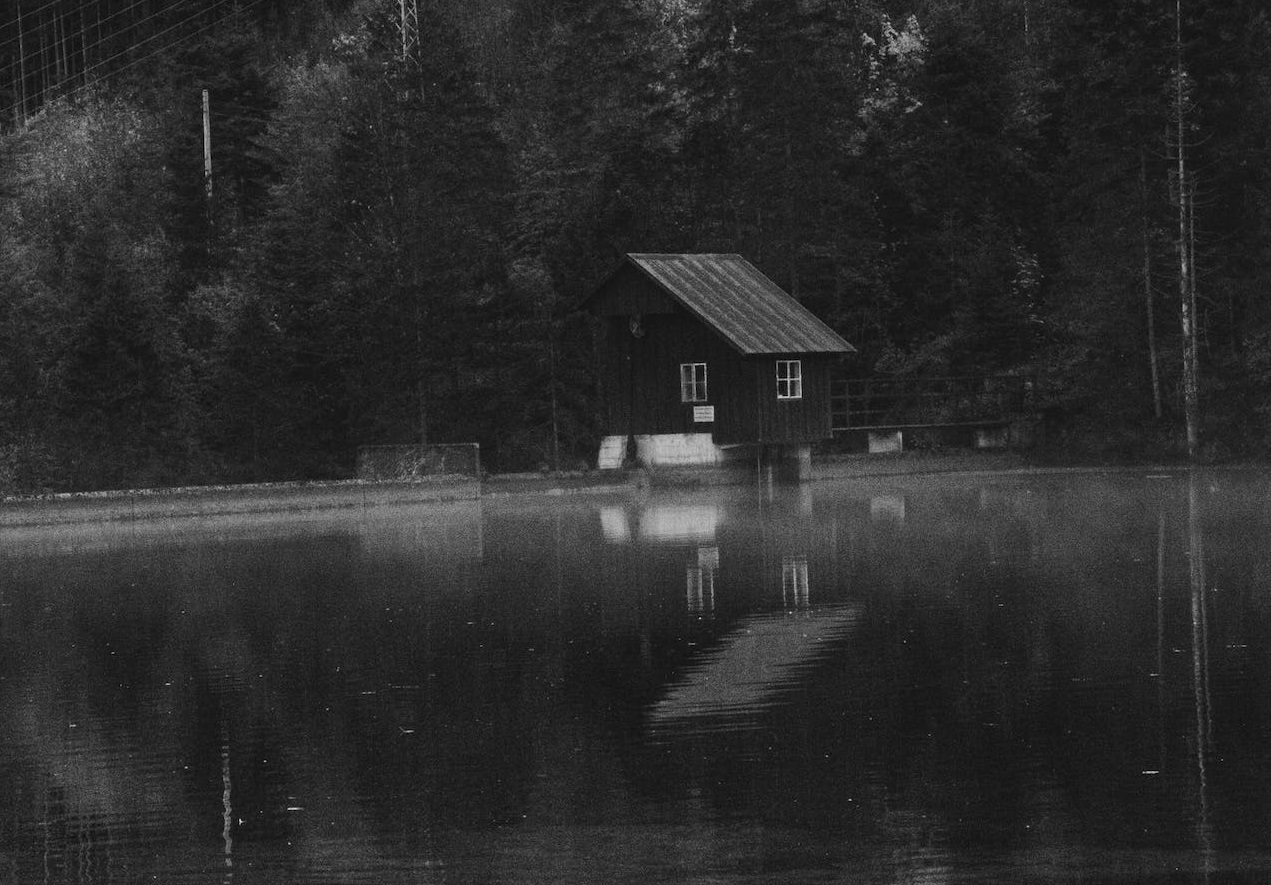 Grayscale photo of house near wood and water.