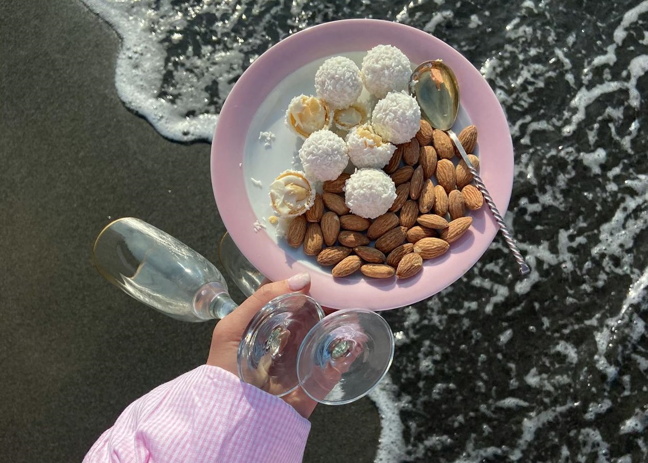 Woman in shirt is walking on the beach with wine glasses and food.